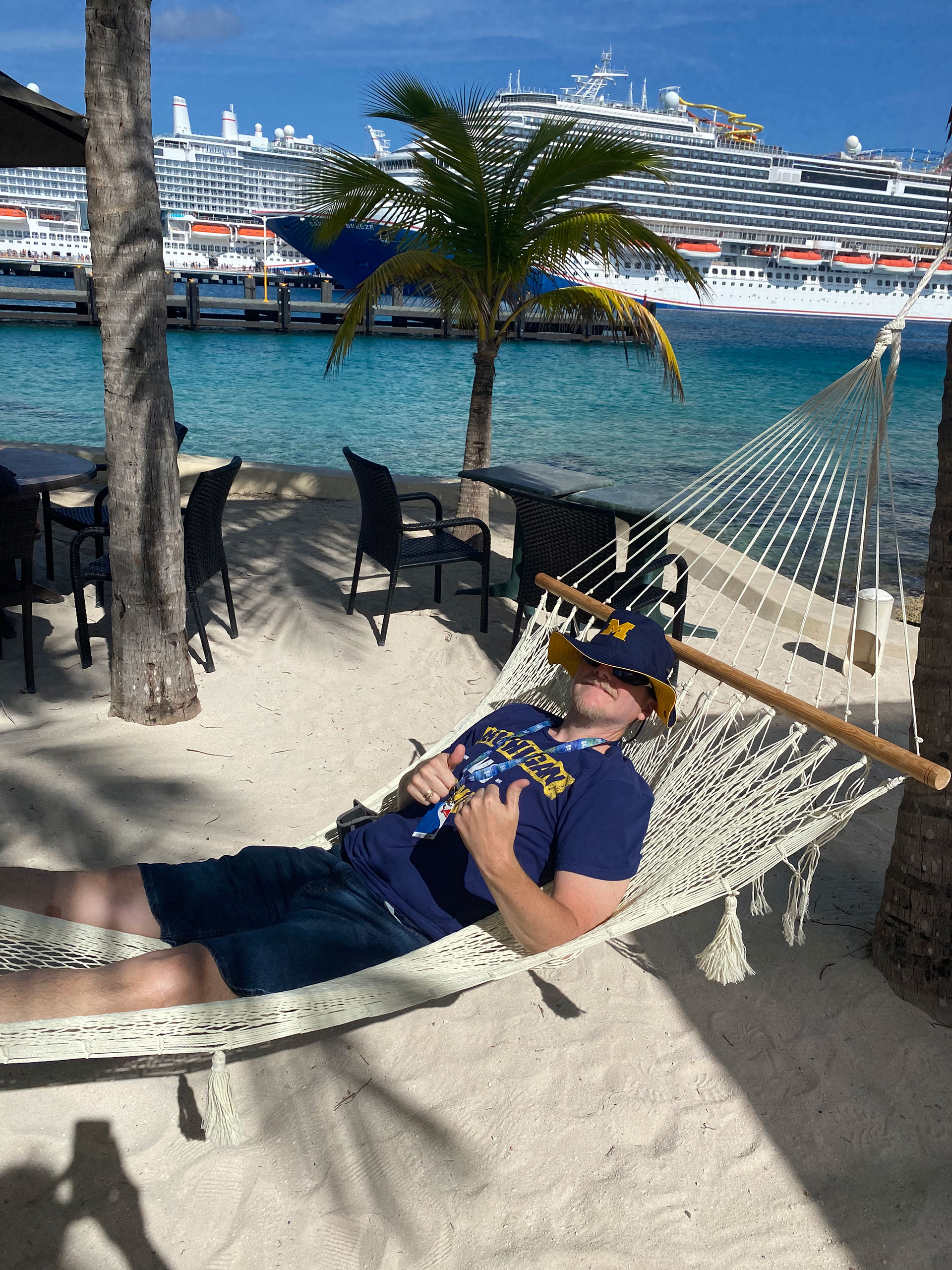 Michigan fan wearing a Michigan t-shirt and Block M bucket hat, laying in a hammock on a beach